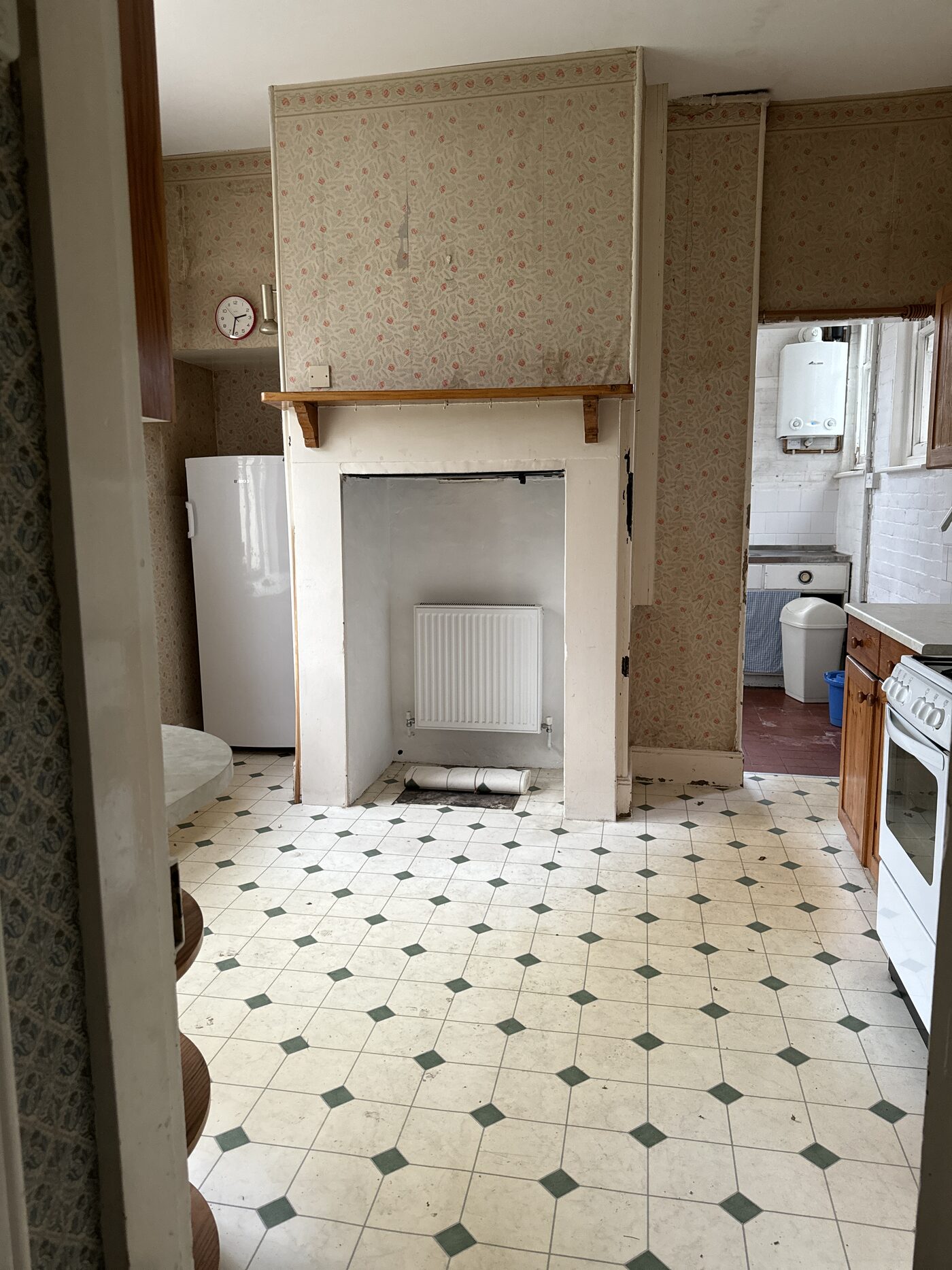 A tired, dated kitchen with old wallpaper, worn flooring and a boxed-in fireplace before renovation